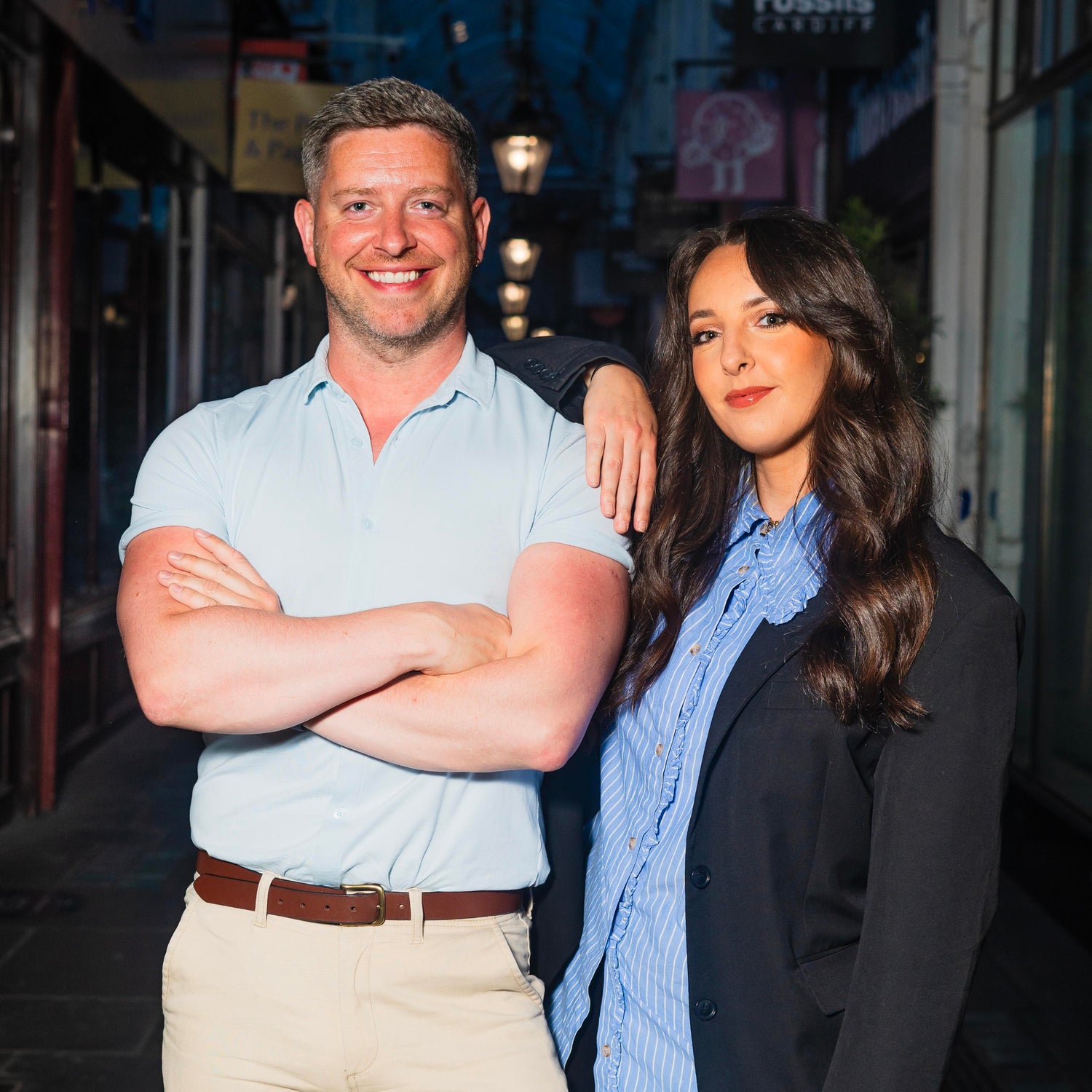 Miriam leans on Geraint's shoulder, as they both stare at the camera in a dimly lit Arcade.