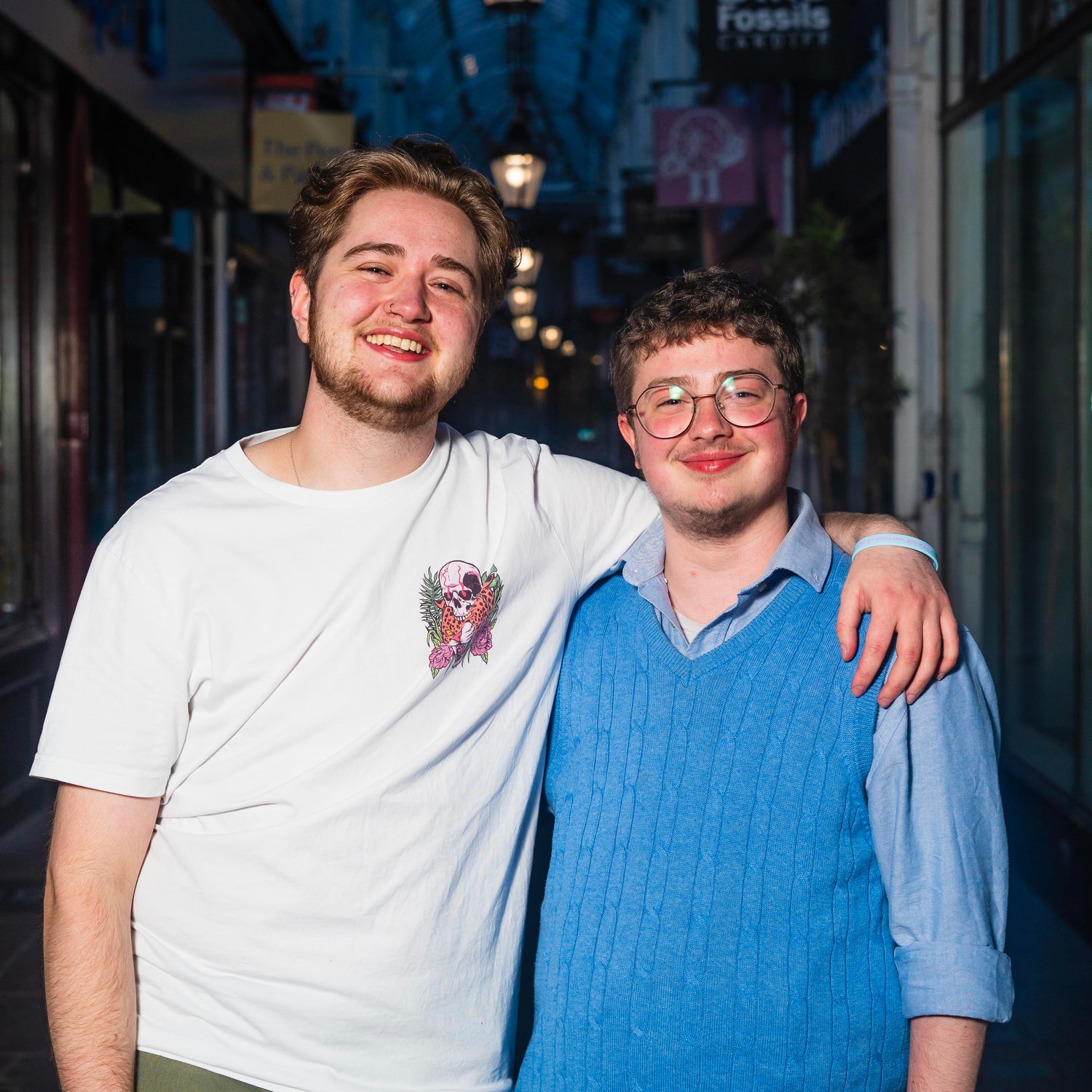Leo and Milo with their arms around each other, smiling at the camera while standing in a dimly lit arcade.